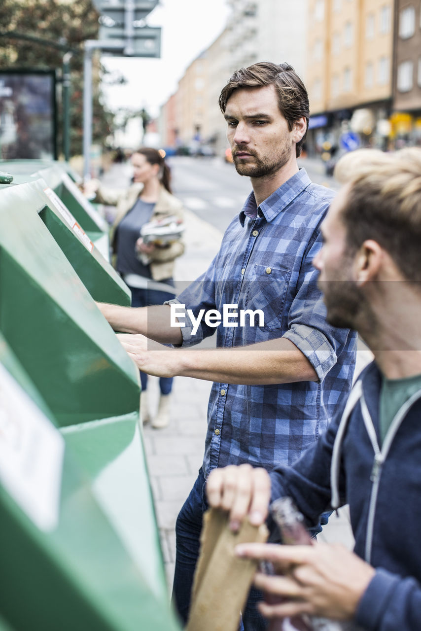 Young man with friends putting recyclable materials into recycling bins