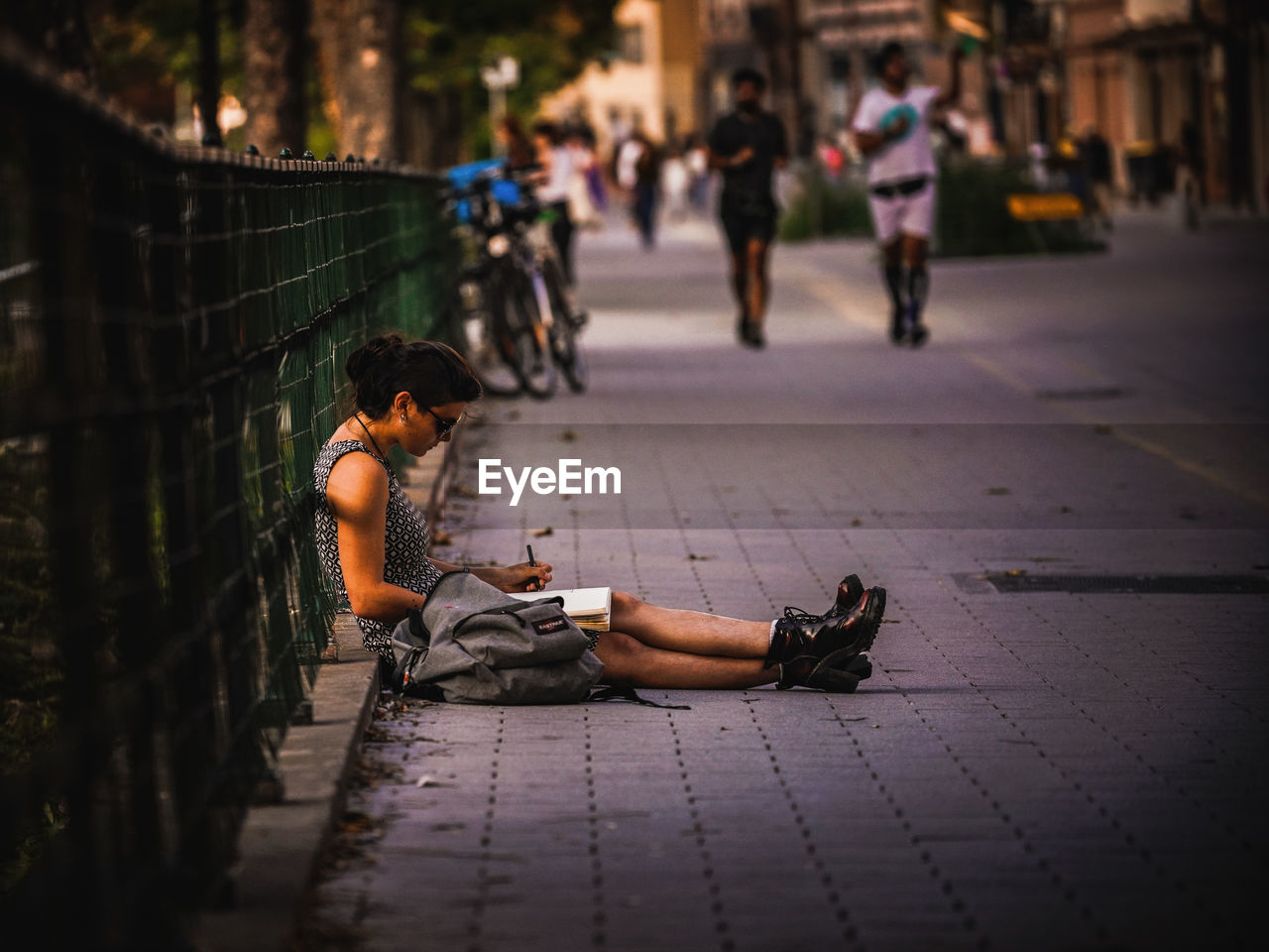 Side view of woman writing in book while sitting on footpath