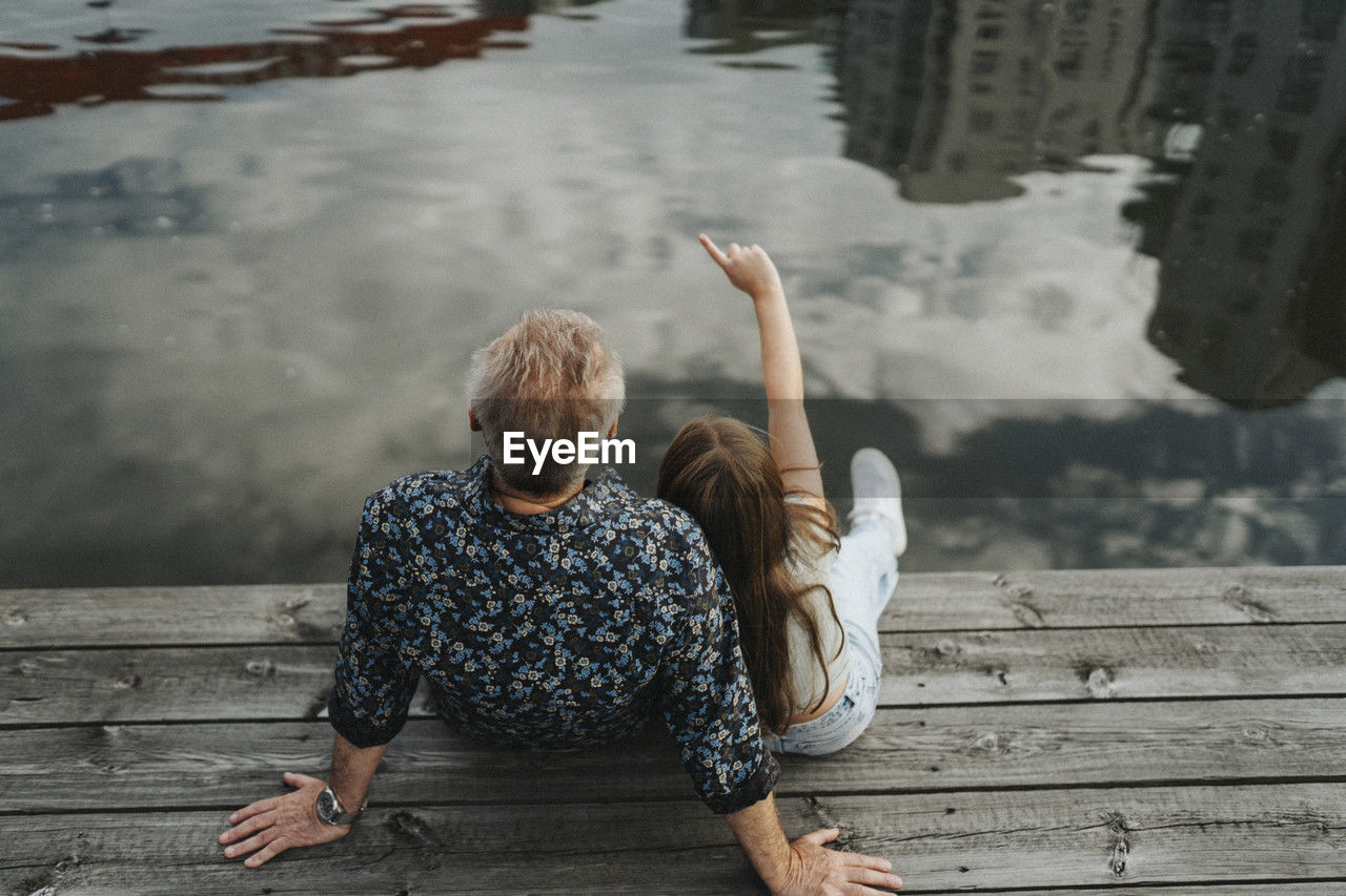 Rear view of girl pointing while sitting with grandfather on jetty