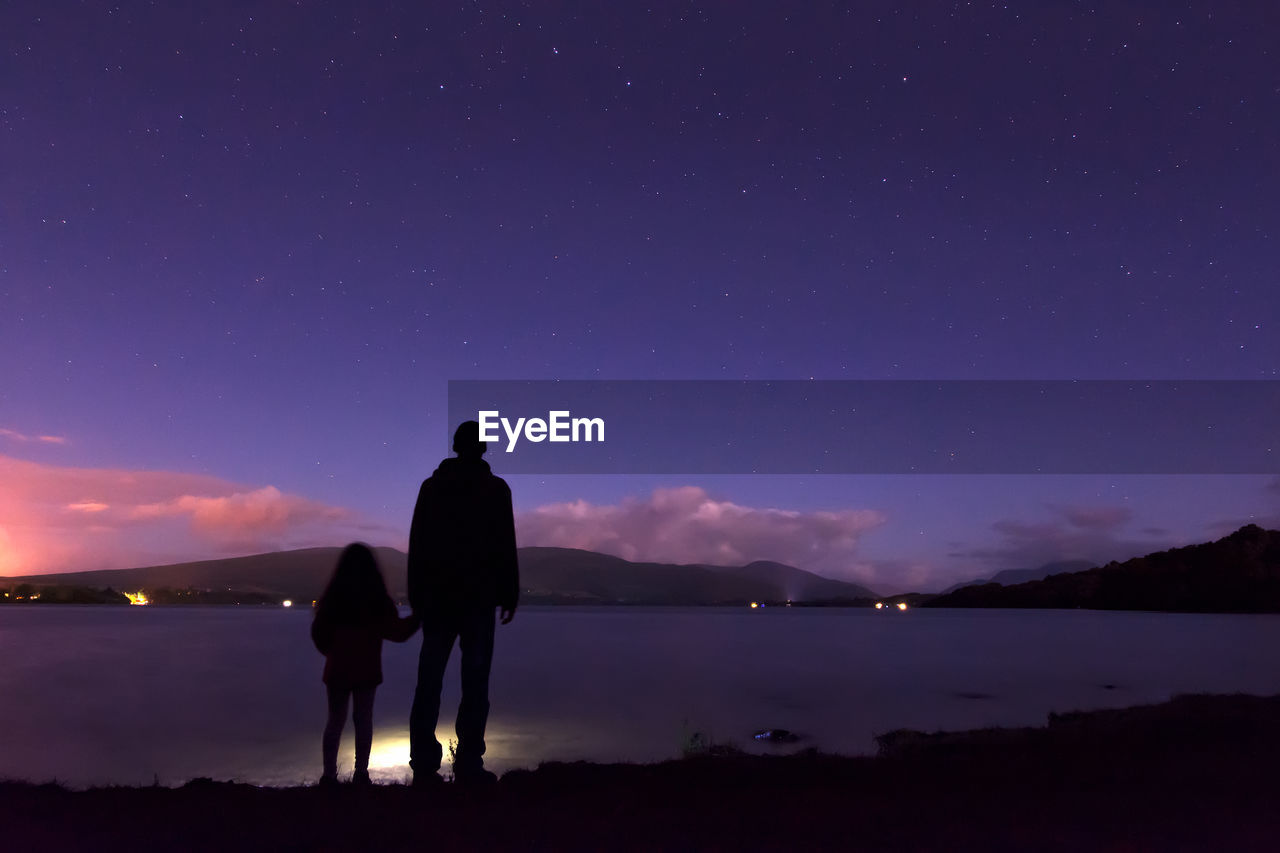 Rear view of father and daughter standing on lakeshore against sky at night