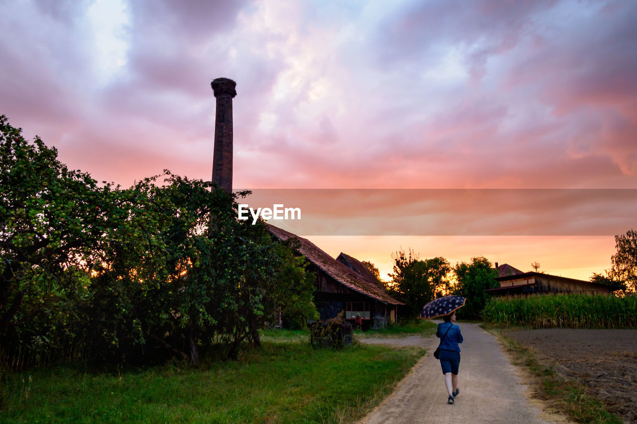 Rear view of mature woman with umbrella standing on road against cloudy sky during sunset