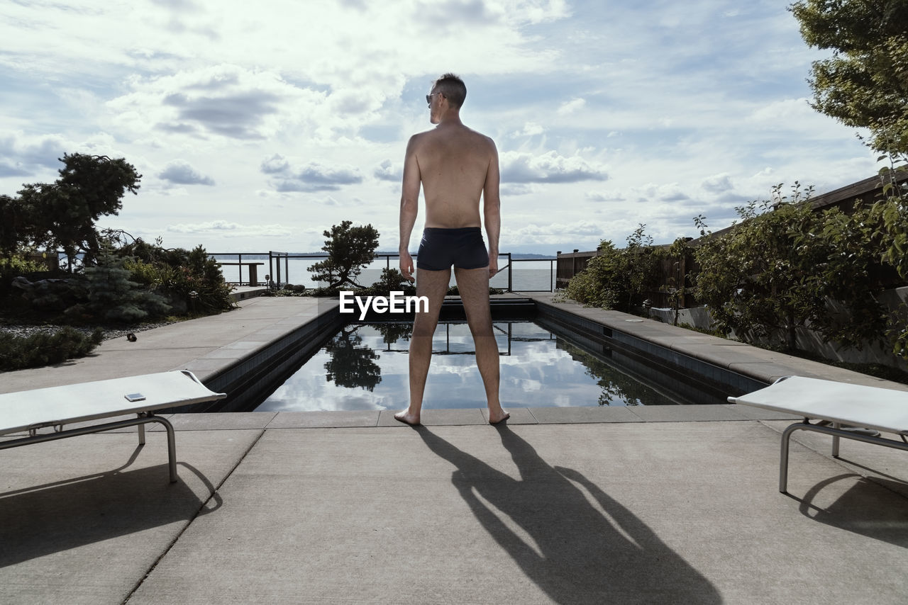 Rear view of shirtless man standing by swimming pool in city against sky