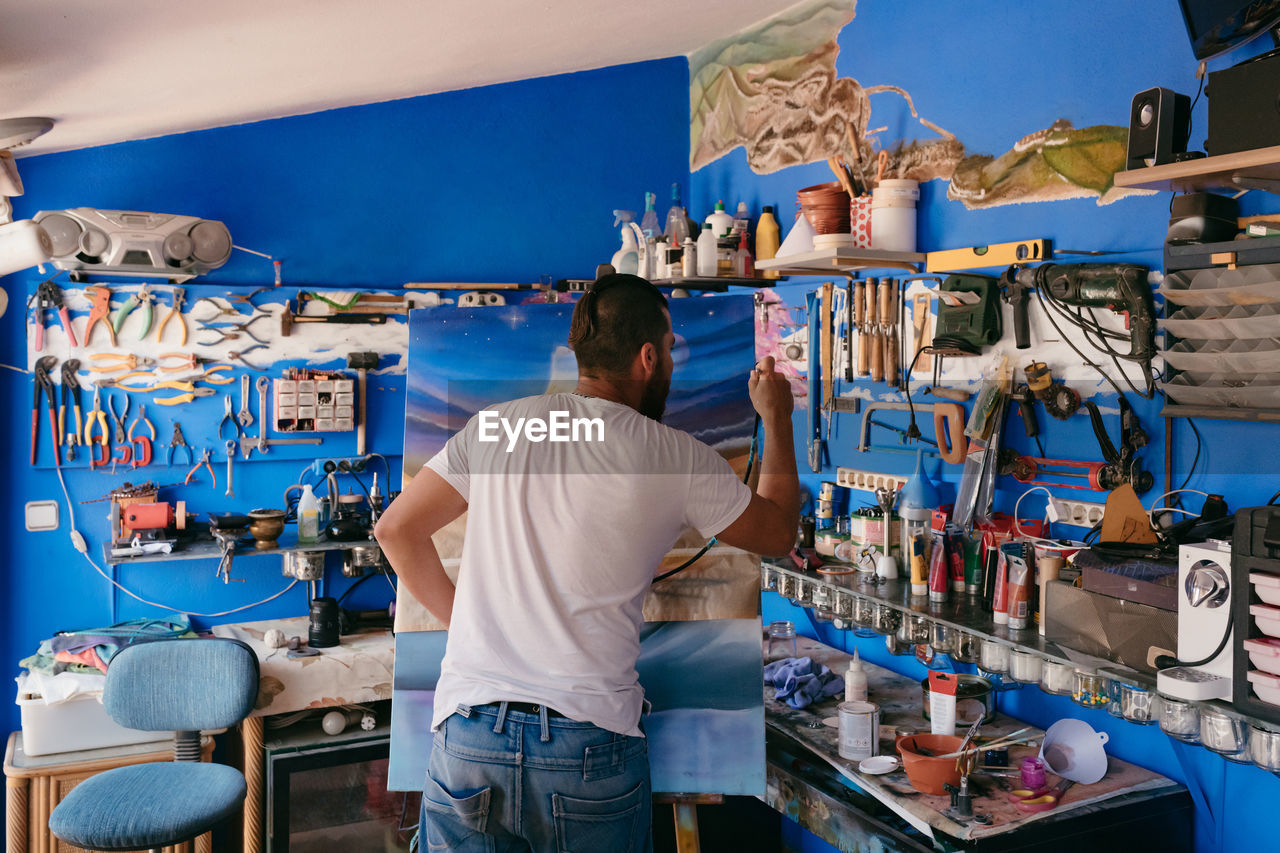 Back view of male artist using spray gun to paint picture on canvas during work in creative workshop