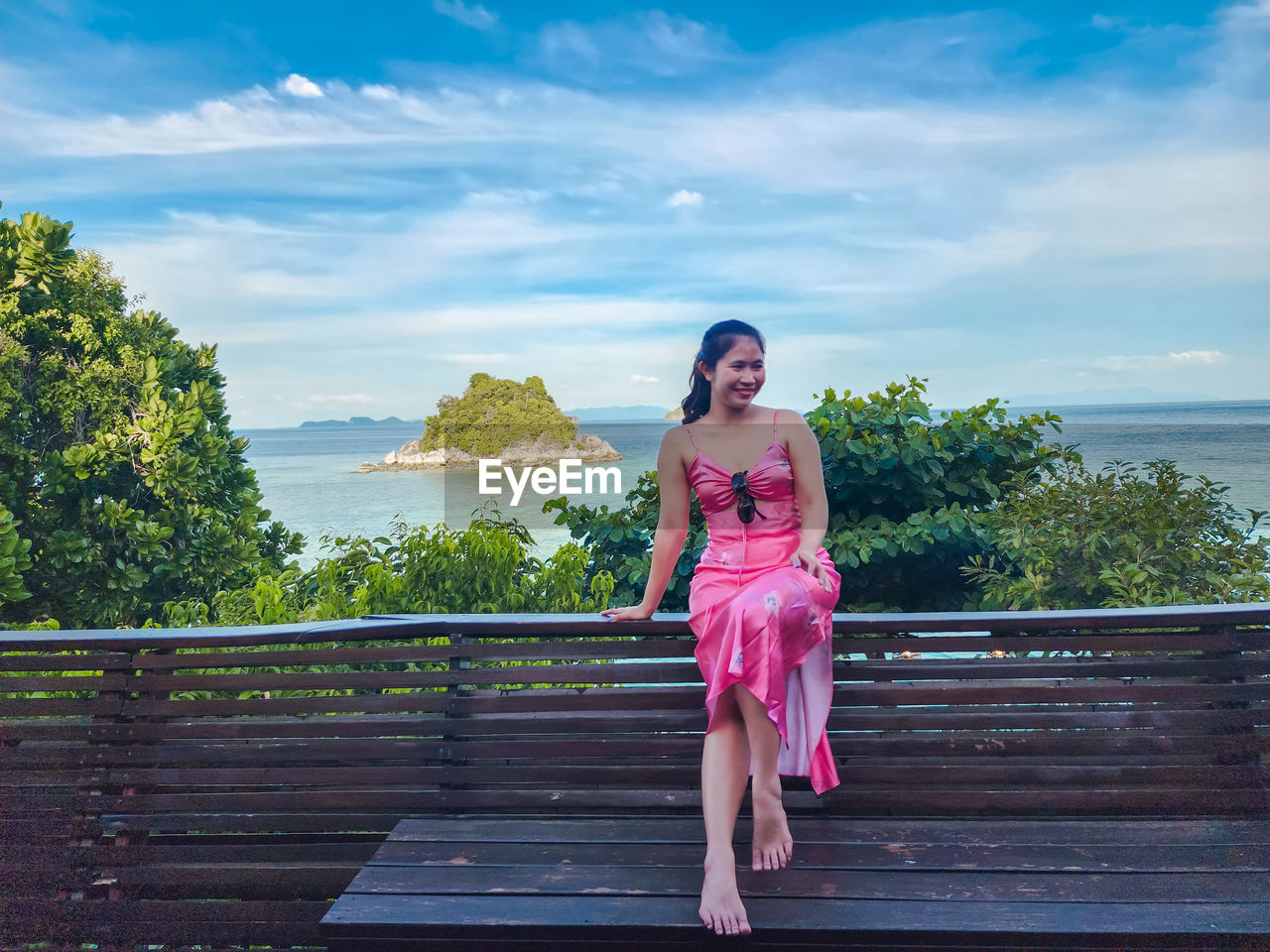one person, women, sky, full length, fashion, vacation, adult, dress, clothing, nature, pink, cloud, young adult, water, hairstyle, lifestyles, female, smiling, beauty in nature, front view, portrait, leisure activity, plant, relaxation, blue, tree, wood, happiness, outdoors, summer, standing, railing, architecture, day, tranquility, sea, long hair, emotion, child, spring, trip, looking at camera, brown hair, looking, land, casual clothing