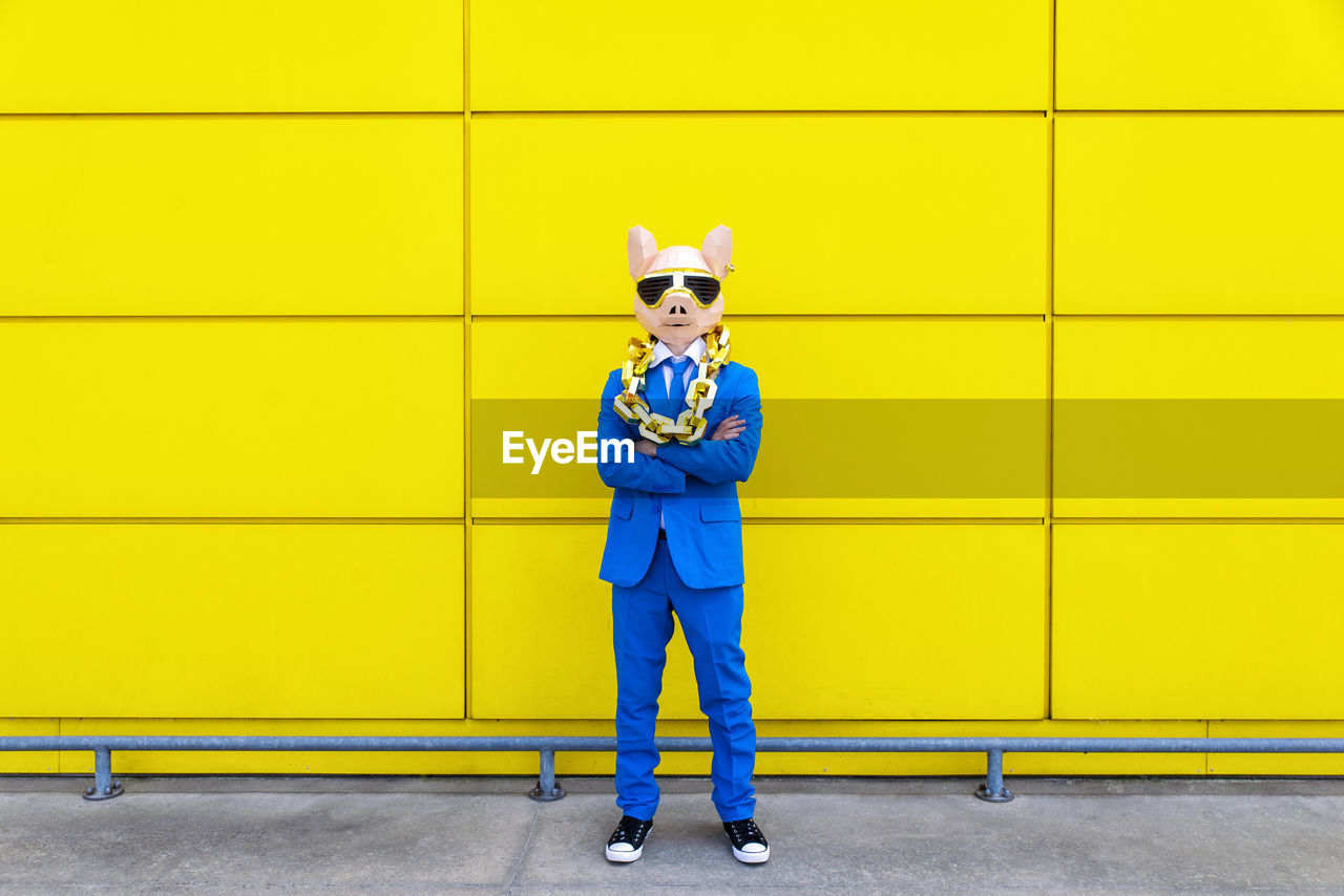 Man wearing vibrant blue suit, pig mask and large golden chain posing against yellow wall