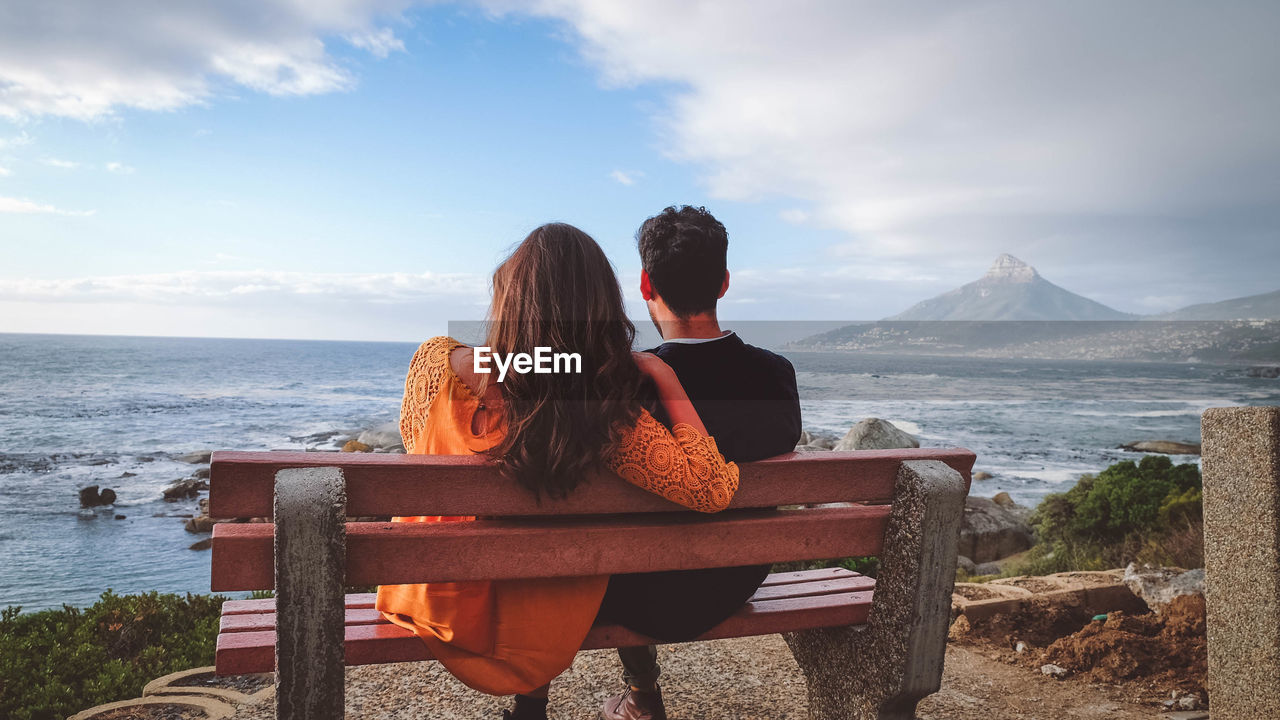 Rear view of couple sitting on bench while looking at sea against sky