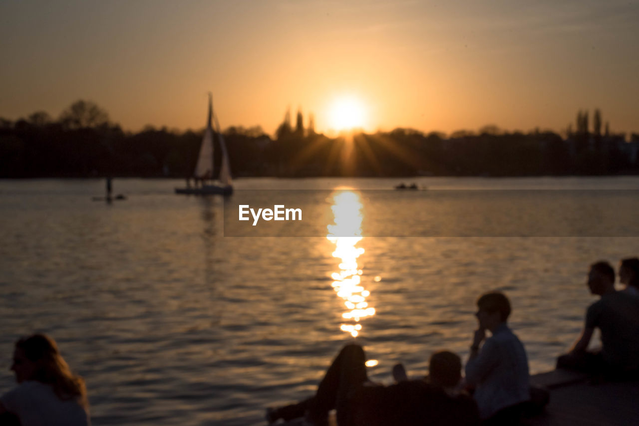 Defocused image of people by lake against sky during sunset