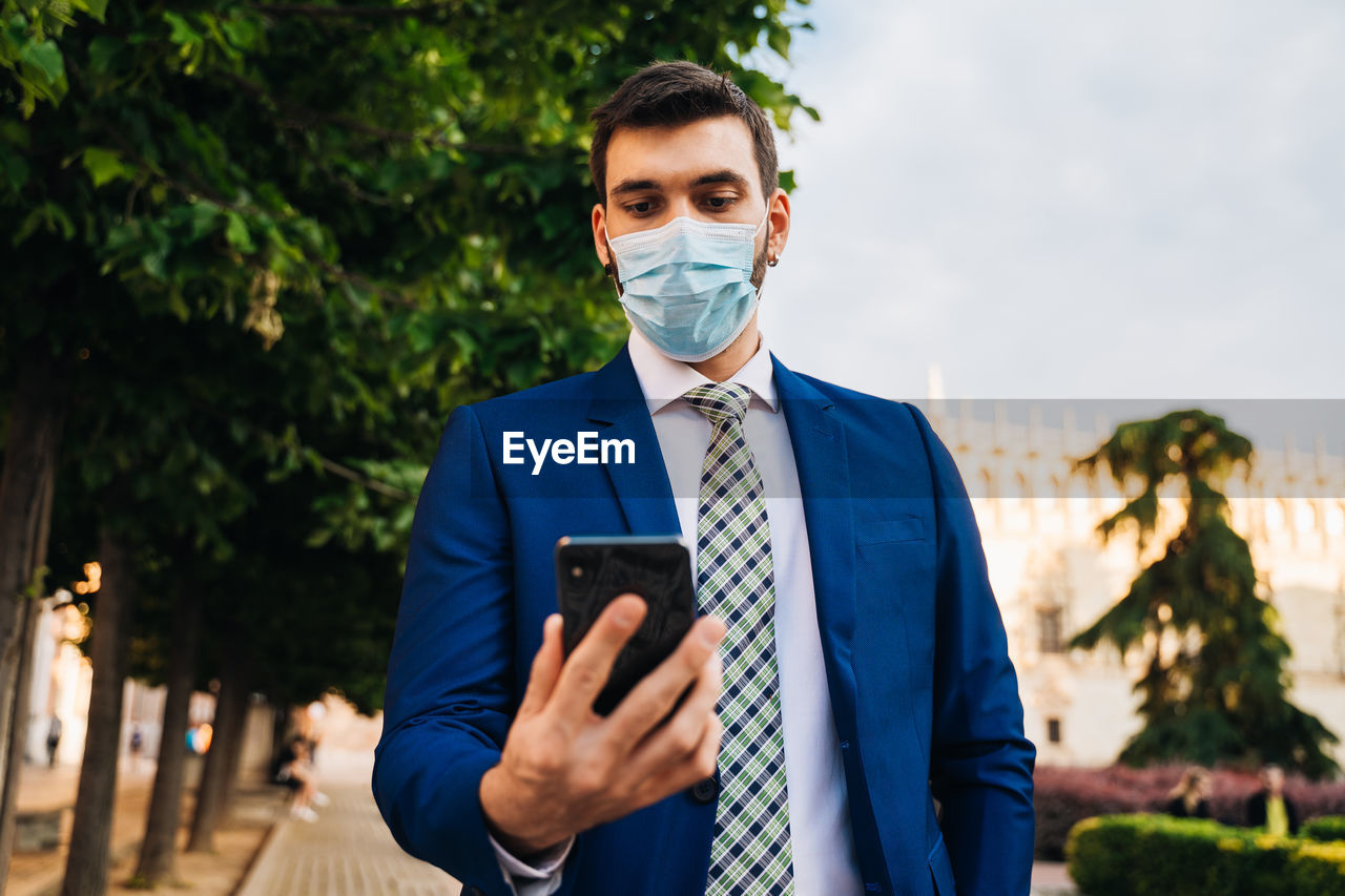 Young elegant businessman with surgical mask looks at phone in park