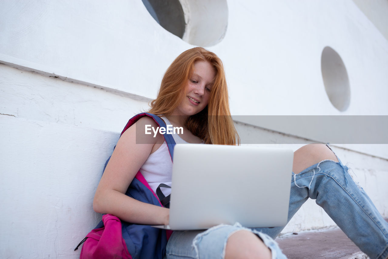 MIDSECTION OF WOMAN USING PHONE WHILE SITTING ON WHITE WALL