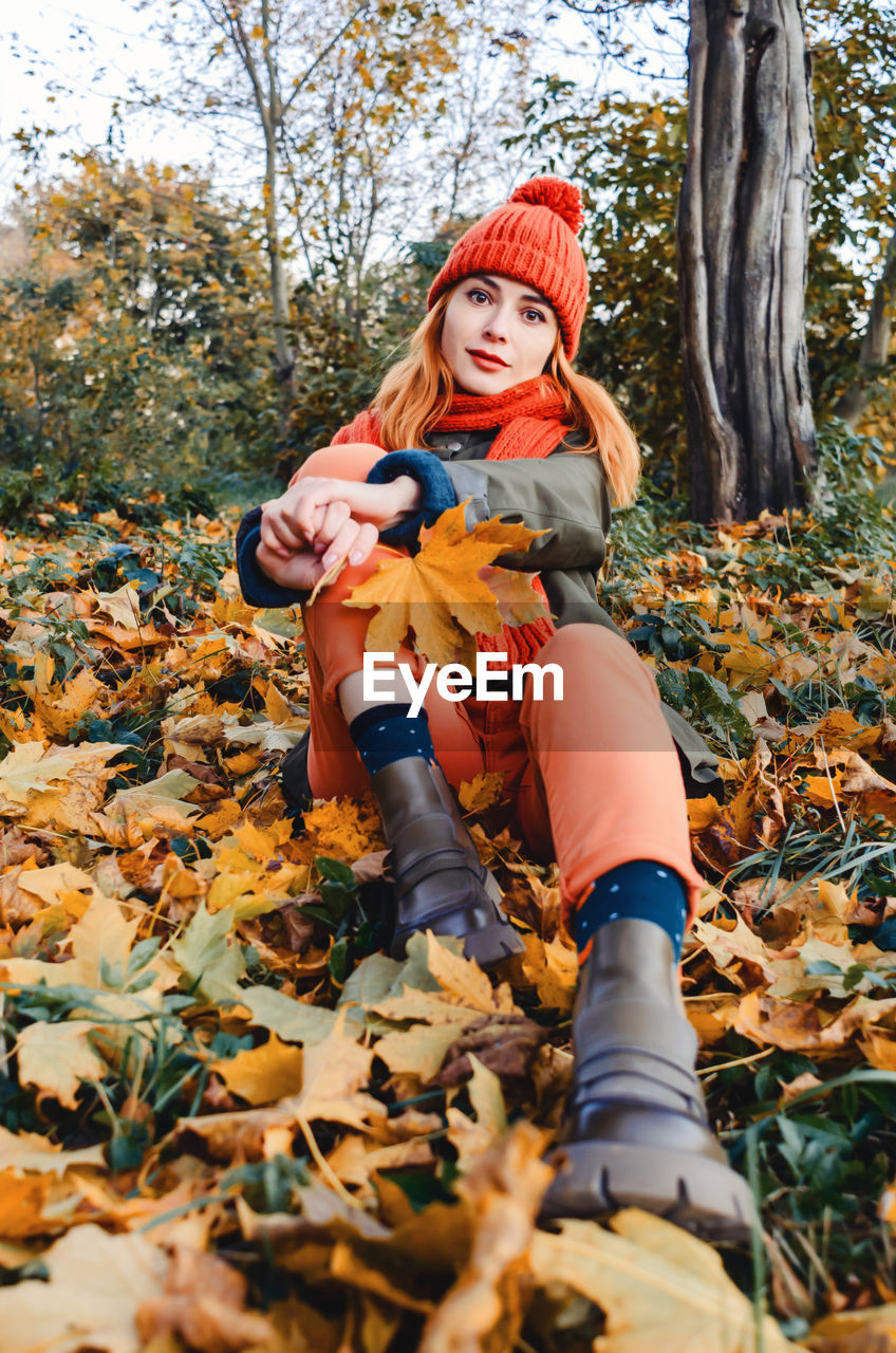 Bright cute young woman in orange warm knitted hat and scarf on background of yellow autumn leaves. 