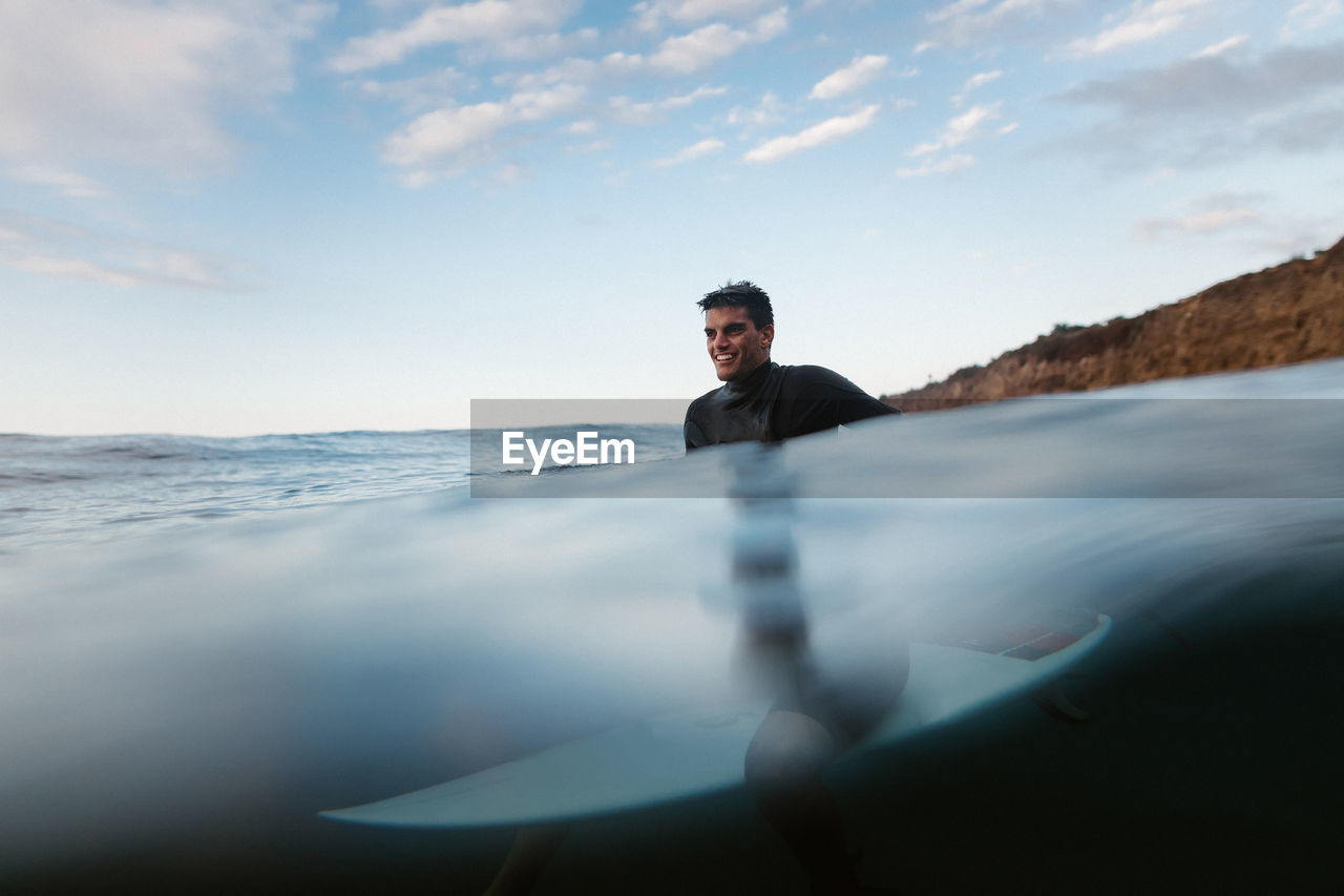 Cheerful male surfer on surfboard in wavy sea looking forward against mount in daytime