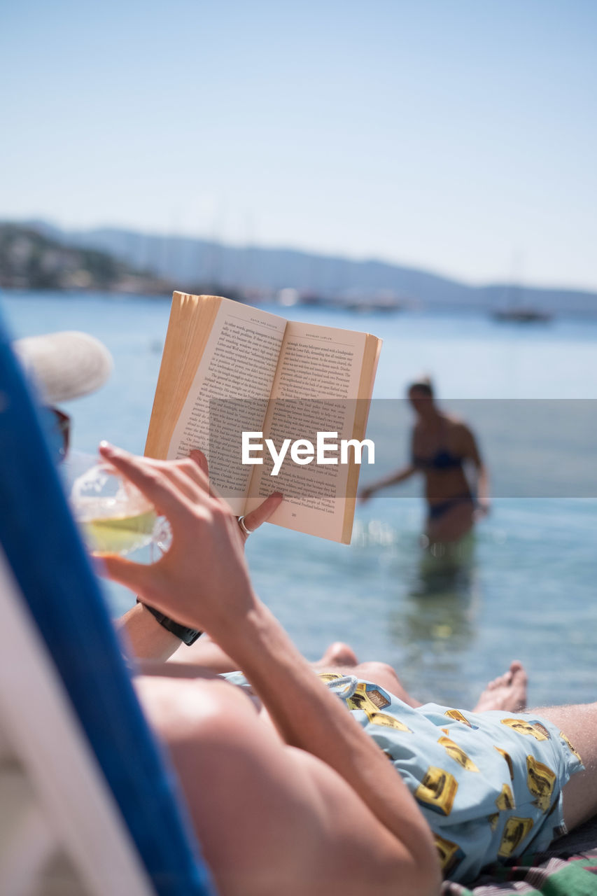 Shirtless man reading book while lying at beach during sunny day