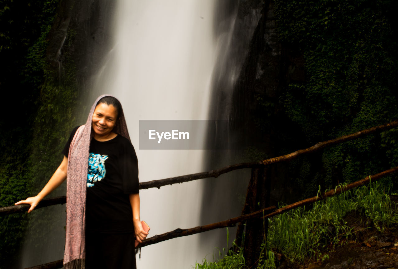 one person, waterfall, young adult, adult, nature, women, sunlight, standing, emotion, water, smiling, darkness, happiness, forest, clothing, lifestyles, land, motion, beauty in nature, outdoors, three quarter length, plant, portrait, front view, railing, casual clothing, leisure activity, tree, hairstyle, light