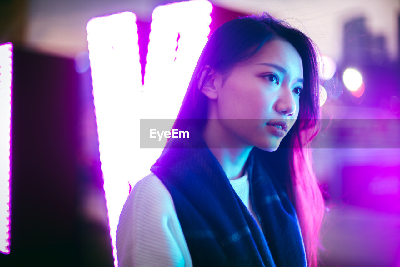 Young woman looking away while standing by illuminated window