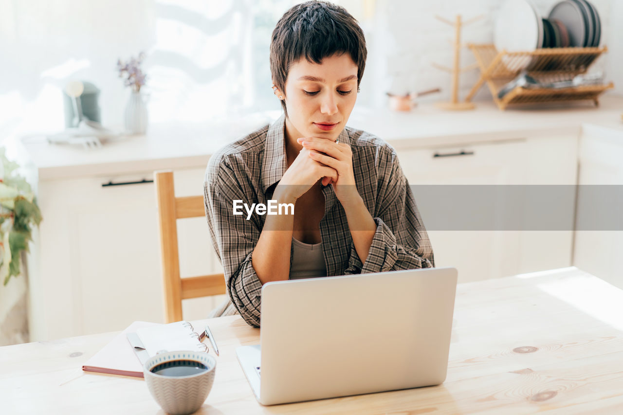 Woman freelancer sitting in the kitchen at home working on a laptop.