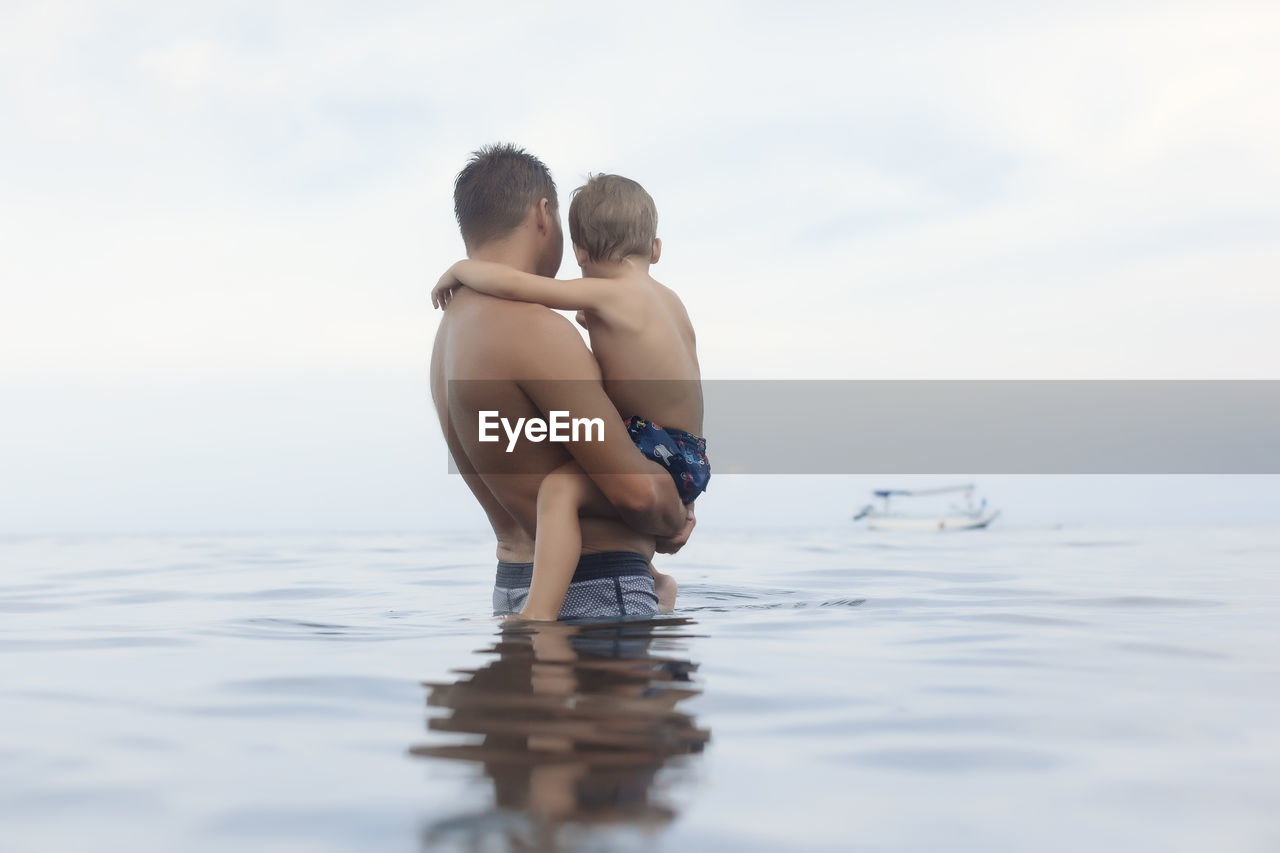 Side view of shirtless father carrying son while standing in sea against cloudy sky