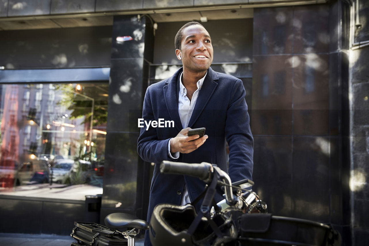 Smiling young male commuter using smart phone while standing with electric bicycle against building in city