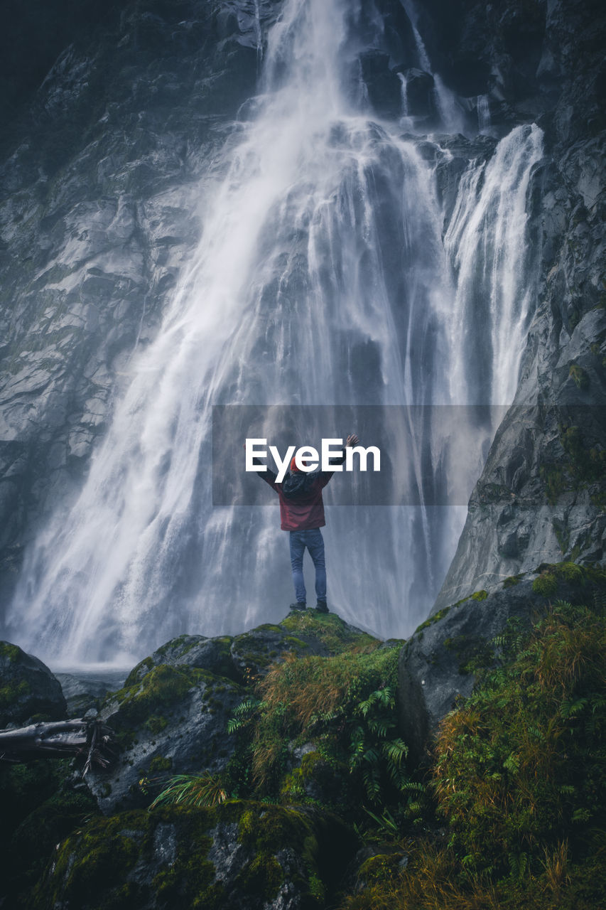 Rear view of man with arms raised standing on rock by waterfall