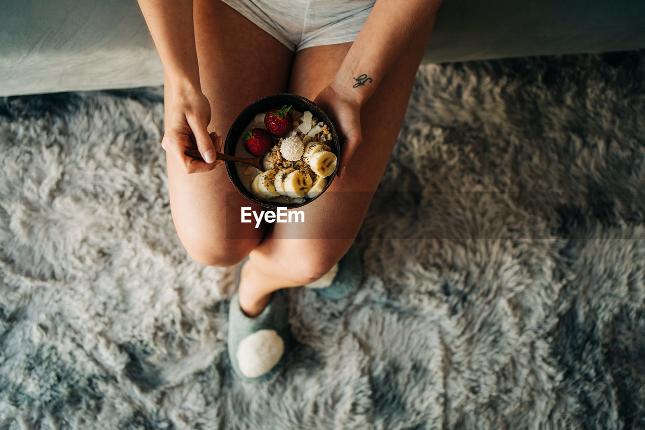 Top view anonymous female in shorts enjoying tasty oatmeal with ripe strawberries and sliced banana while sitting on bed