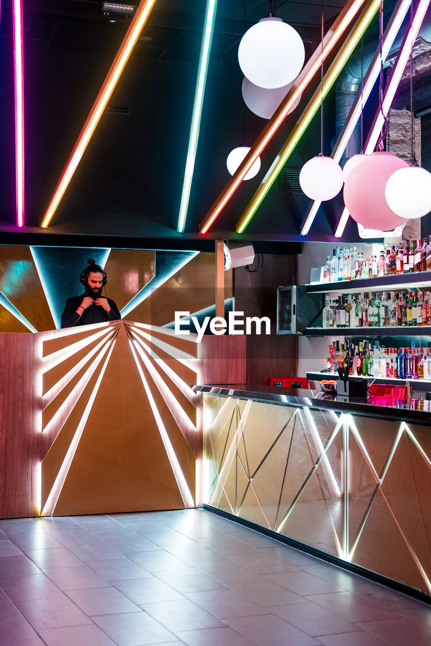 Concentrated young bearded male dj in headphones standing at illuminated counter in modern nightclub