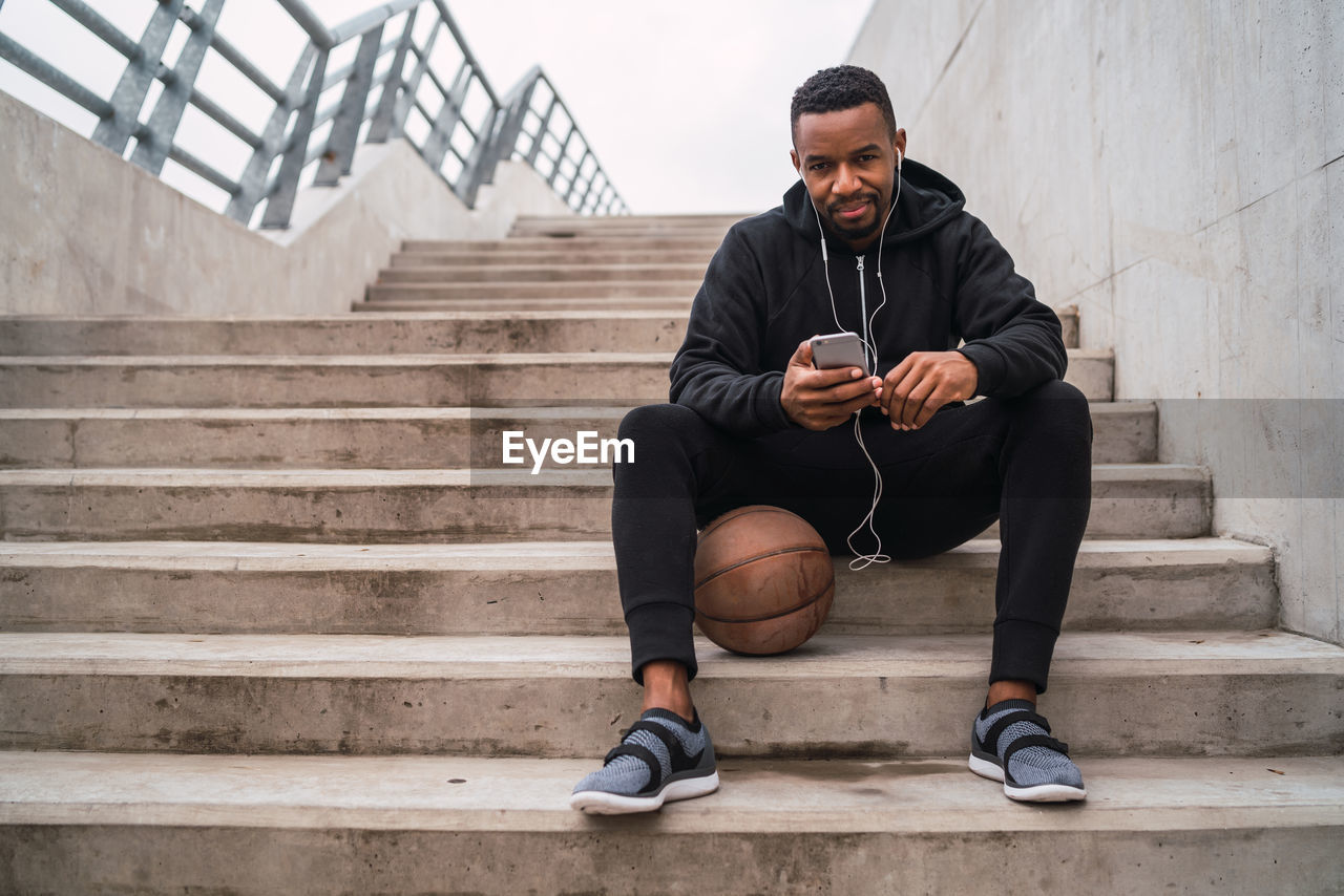 Portrait of man listening music while sitting on steps with basketball