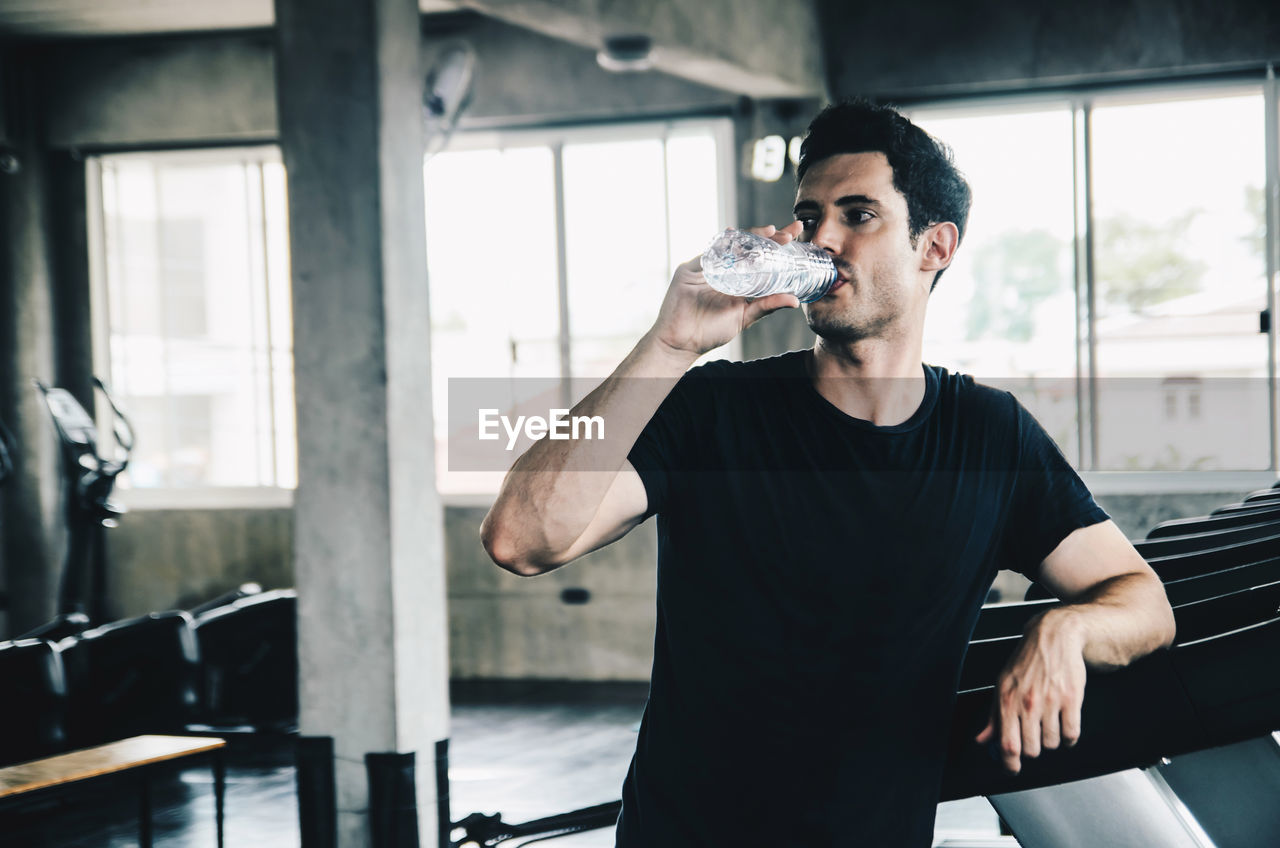 Thoughtful young man drinking water while standing in gym