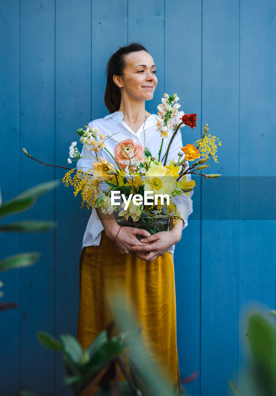 Woman in flowing yellow skirt and white shirt with bouquet in glass vase against the blue background