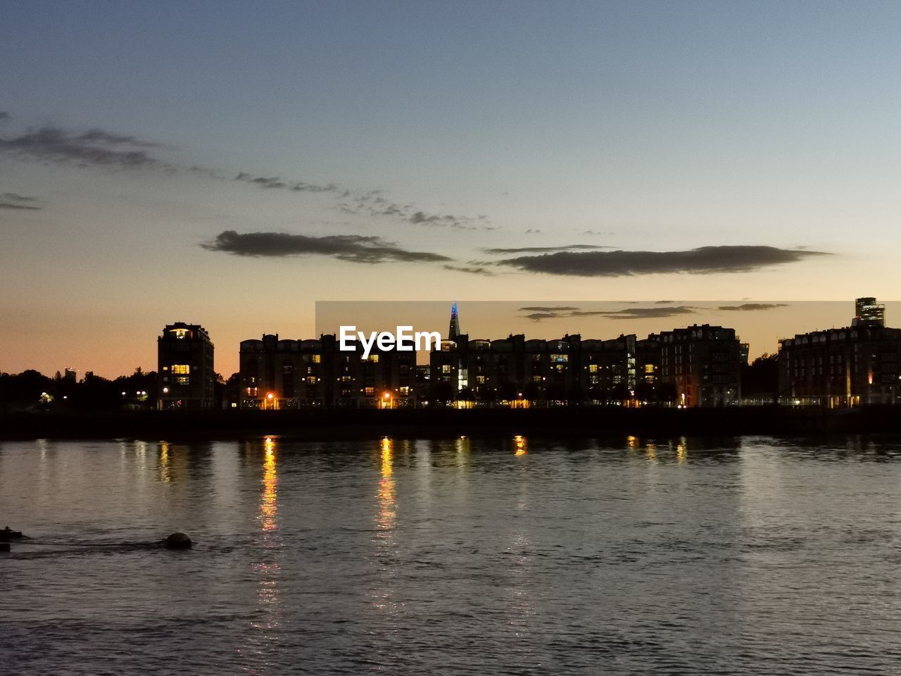 RIVER BY ILLUMINATED CITY AGAINST SKY AT DUSK