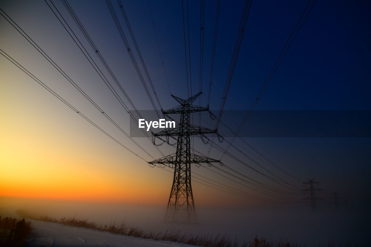 Low angle view of electricity pylon against sky during sunset, standing in a foggy ground