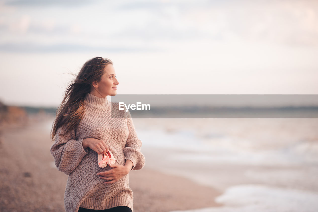 Smiling pregnant woman wear knit cardigan hold baby shoes posing over sea background close up.