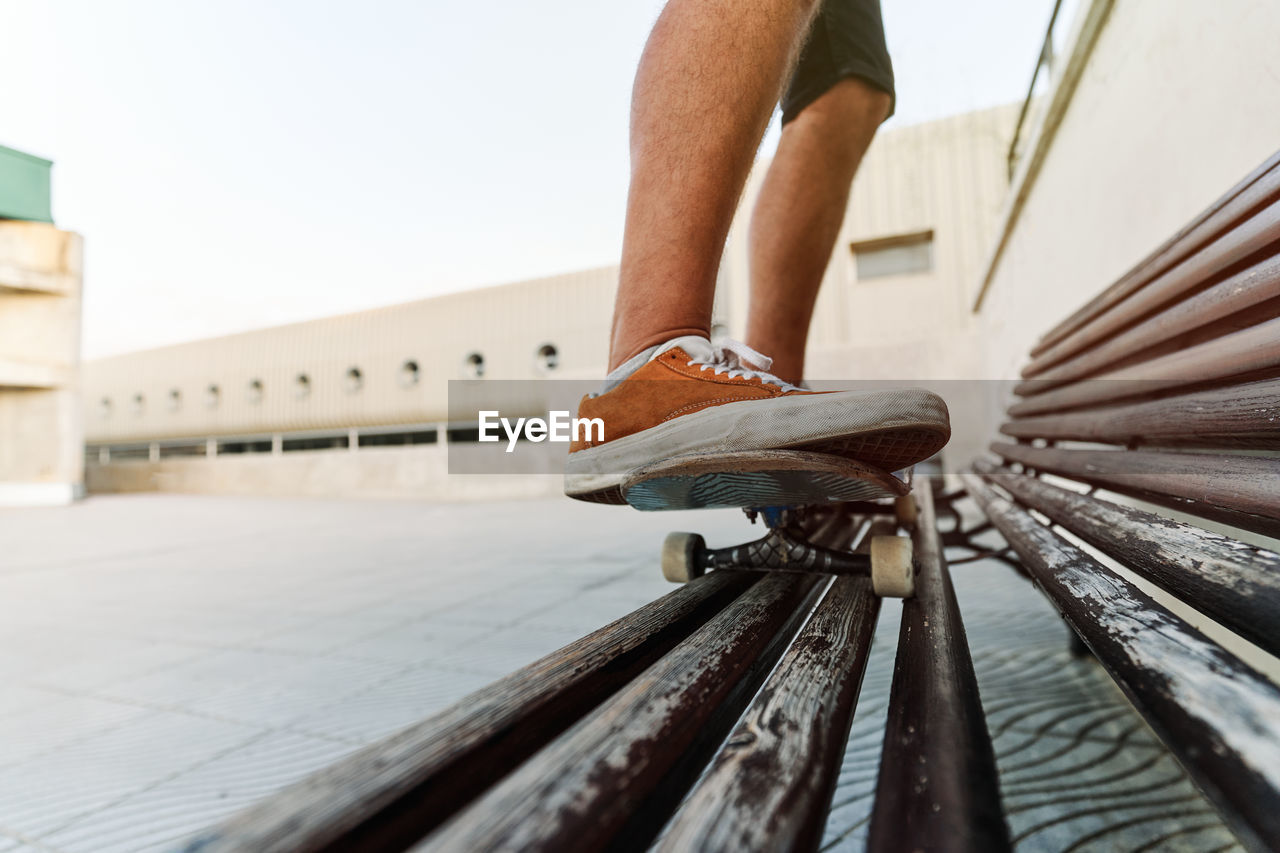 Low angle of unrecognizable crop male standing on skateboard on wooden bench and showing stunt