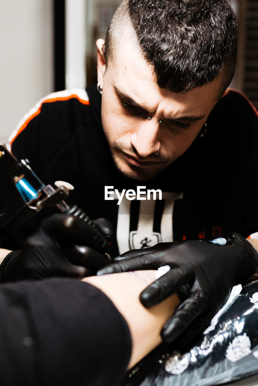 Stylish man with piercing using tattoo machine to make tattoo on leg of crop customer during work in salon