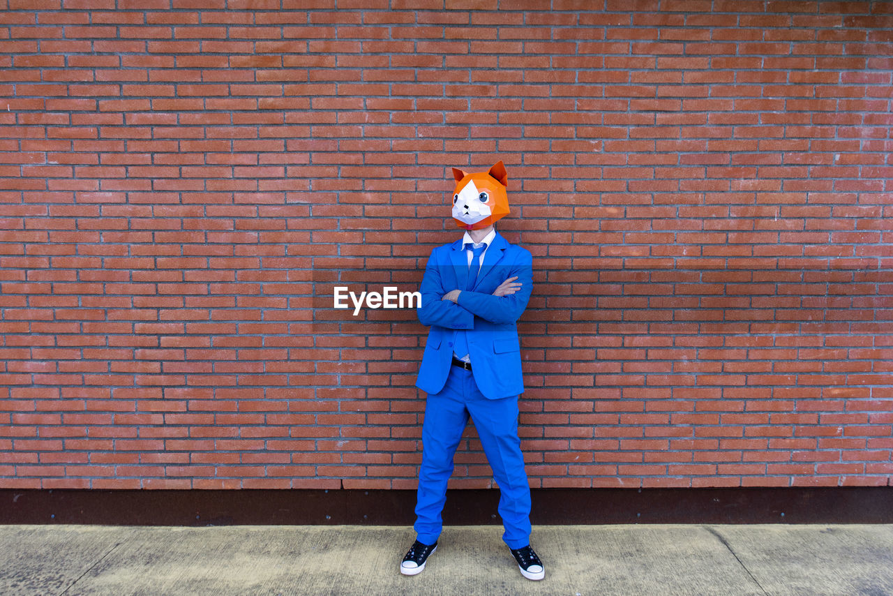 Man wearing vibrant blue suit and cat mask standing with crossed arms in front of brick wall