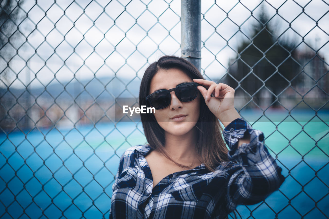 Beautiful young woman wearing sunglasses leaning on chainlink fence