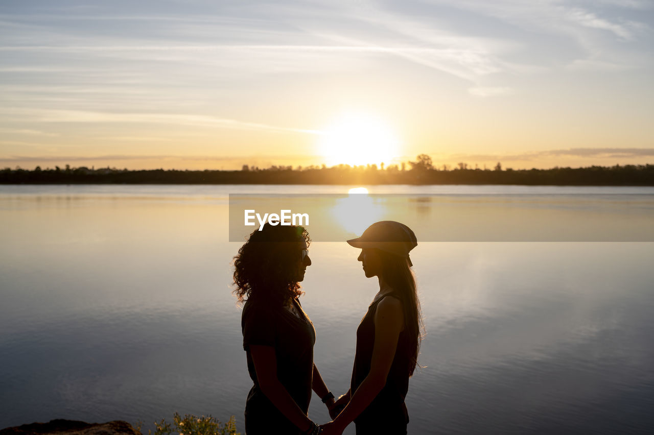 Lesbian couple looking at each other and hugging while standing by a lake at sunset.