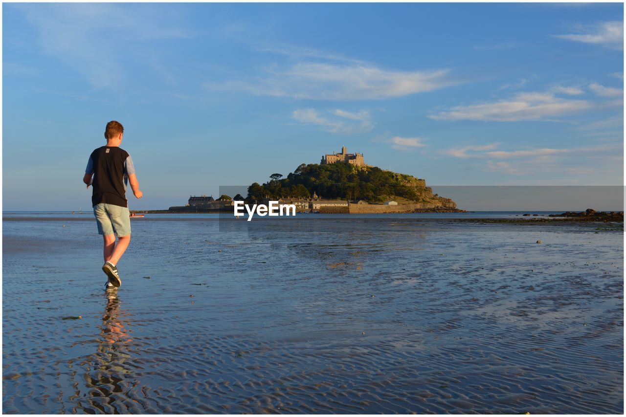 Rear view of young man standing on beach against sky in front of st michaels mount