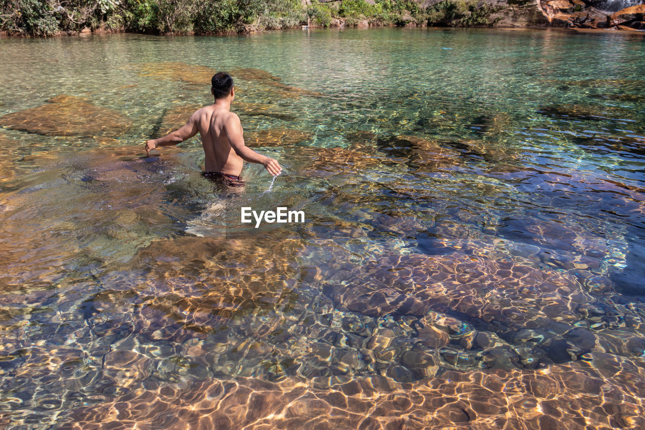 Young man wearing sun glass sitting on rock in flowing river clear water at morning from flat angle