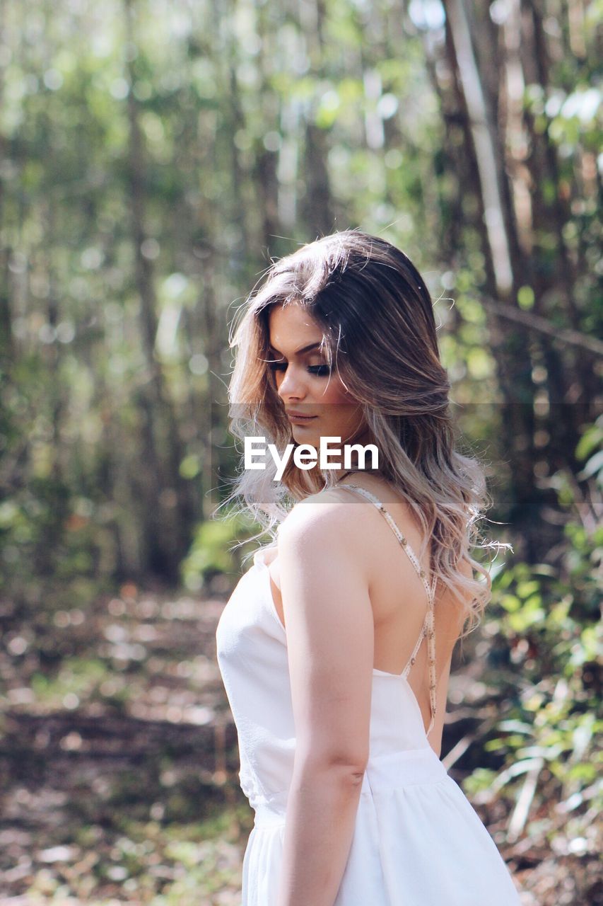 Side view of young woman standing in forest on sunny day