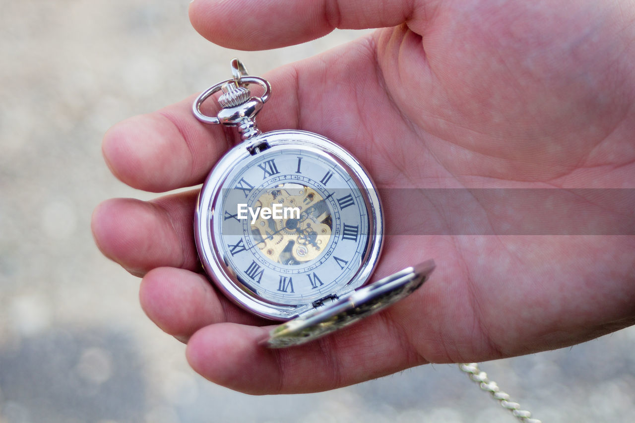 CLOSE-UP OF HUMAN HAND HOLDING CLOCK AGAINST BLURRED BACKGROUND