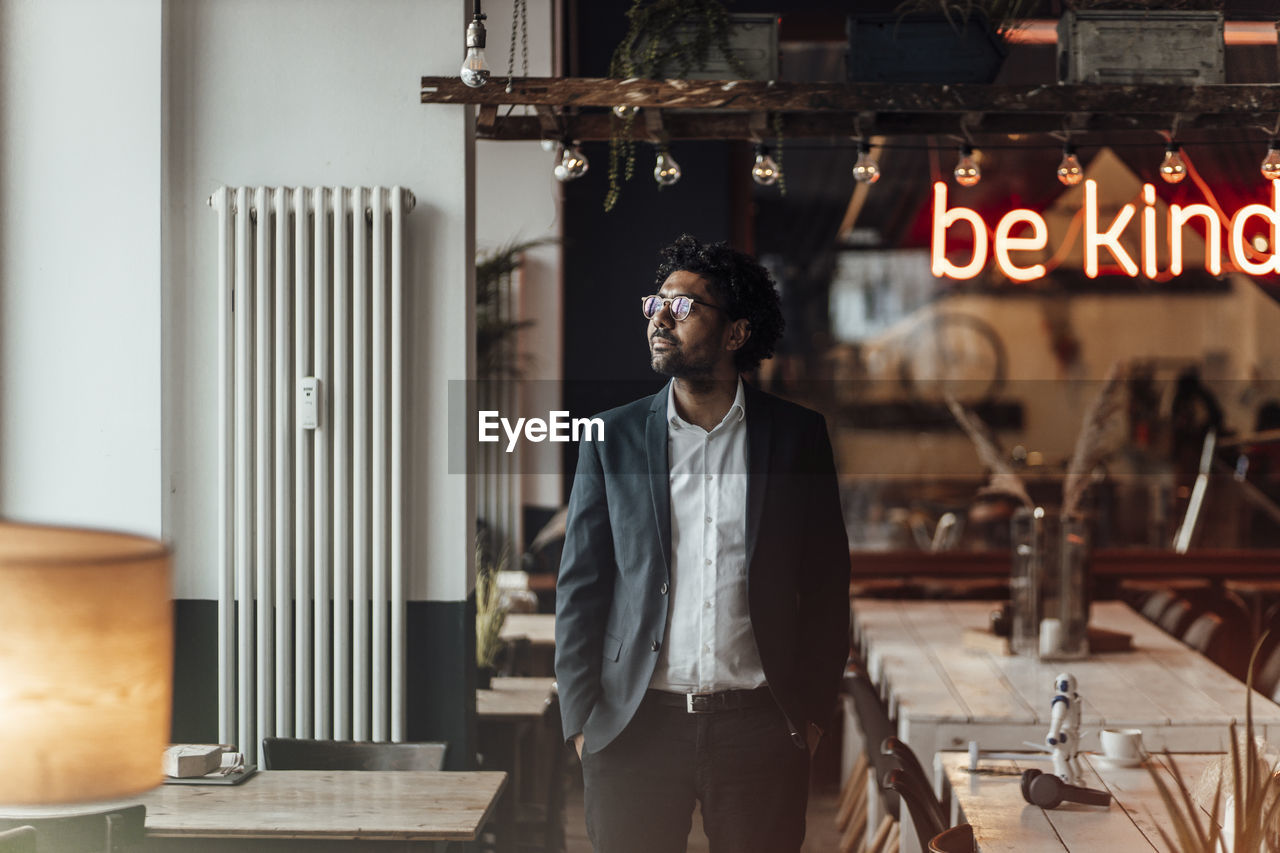 Male professional looking away while standing with hands in pockets at cafe