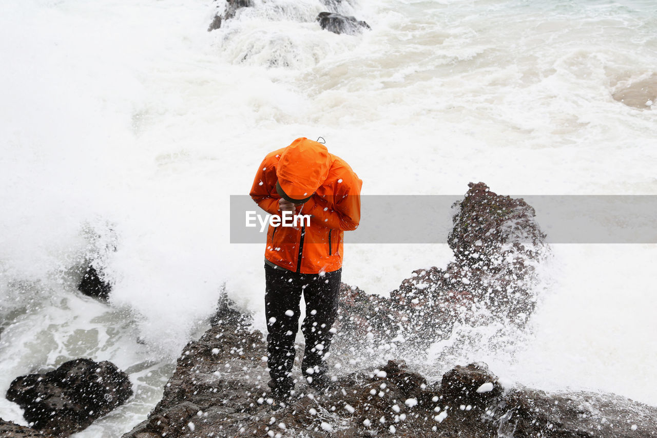 Man in waterproof coat standing on rocks at beach splashed by waves