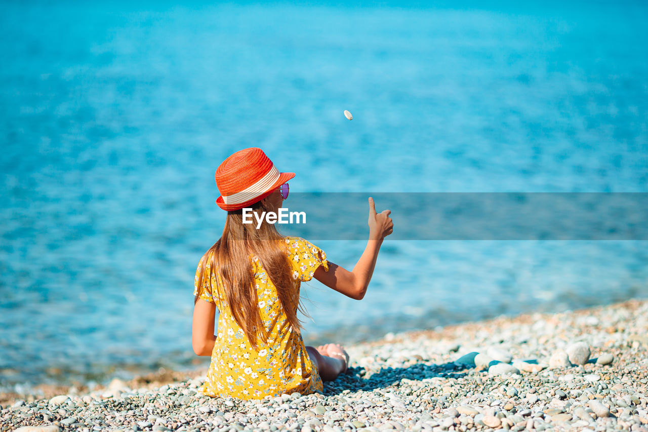 REAR VIEW OF WOMAN SITTING ON ROCK AT BEACH