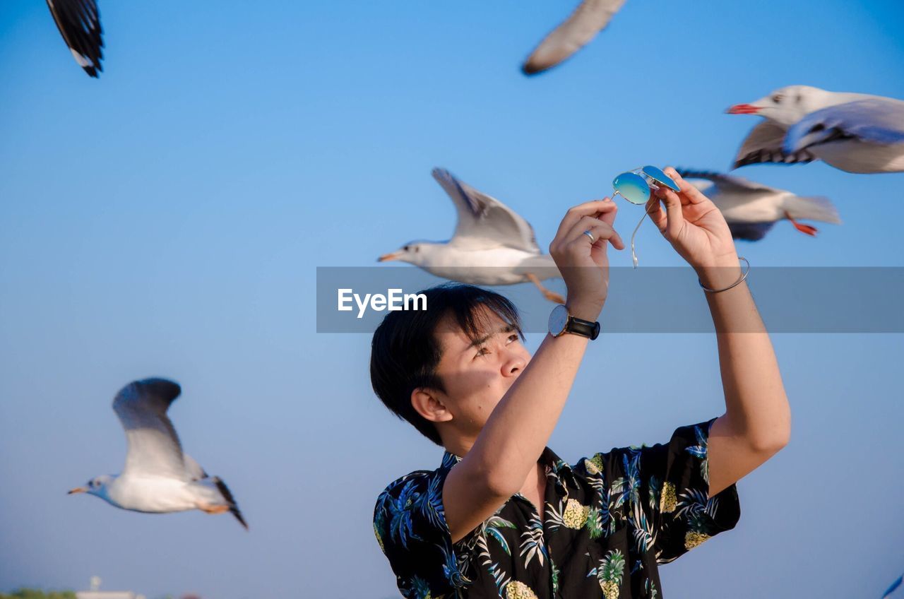 Man holding sunglasses while standing by seagulls flying against sky