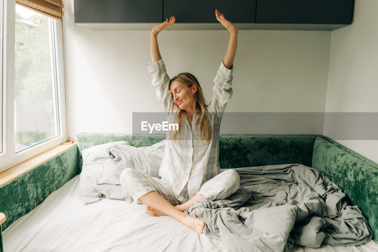 Portrait of beautiful young woman in pajamas sitting on bed, stretching after waking up