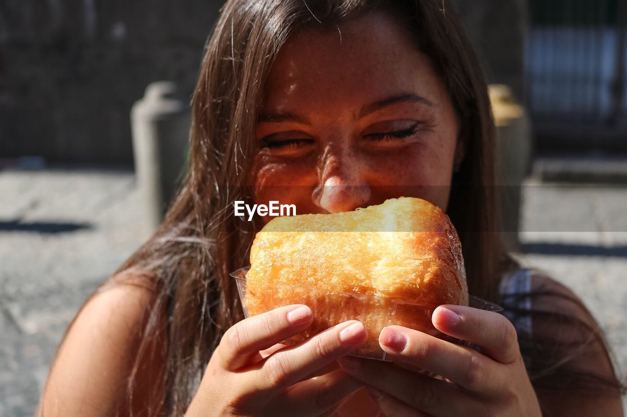 Happy woman eating pastry while standing on city street during sunny day