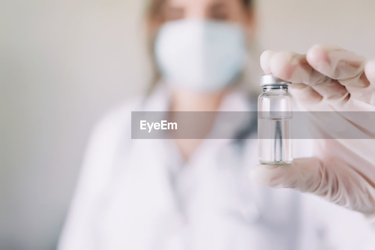 Doctor or nurse in uniform, gloves, face mask in lab, holding medicine liquid vaccine vial bottle