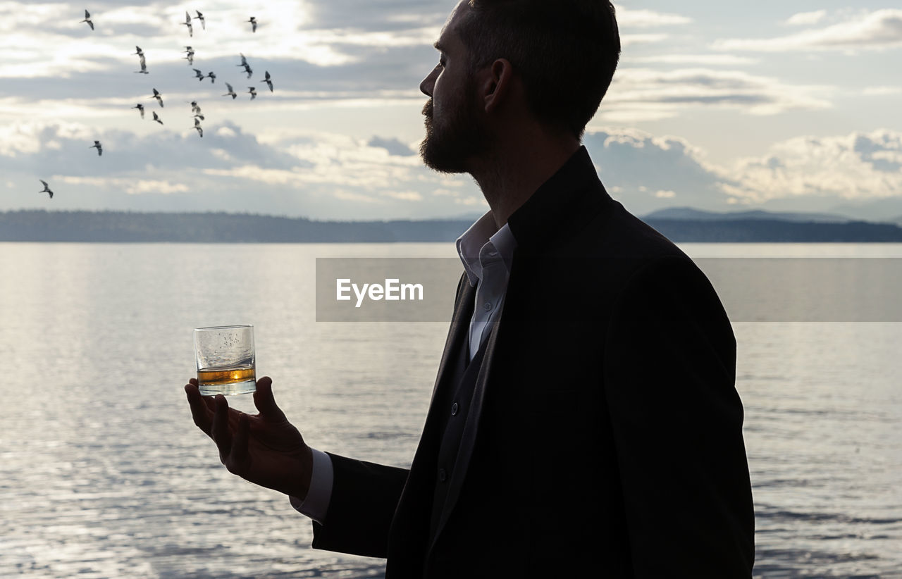 Man holding whiskey glass while standing against sea during sunset