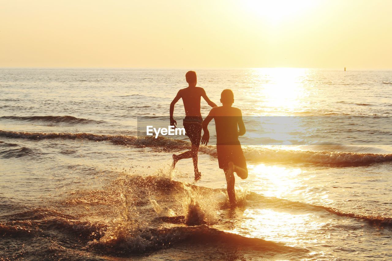 Rear view of silhouette friends running in sea against sky during sunset