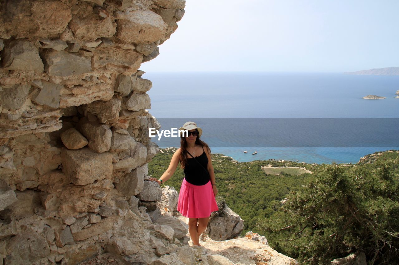 Young woman wearing sunglasses standing on cliff against sea