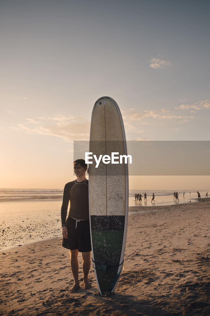 Full length of young man with surfboard standing at beach during sunset